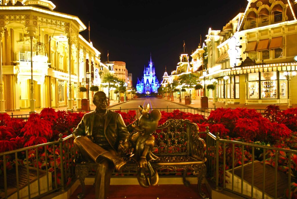 Walt Disney with Minnie Mouse, sitting on a bench in front of a lit up Main Street-Disney World at night