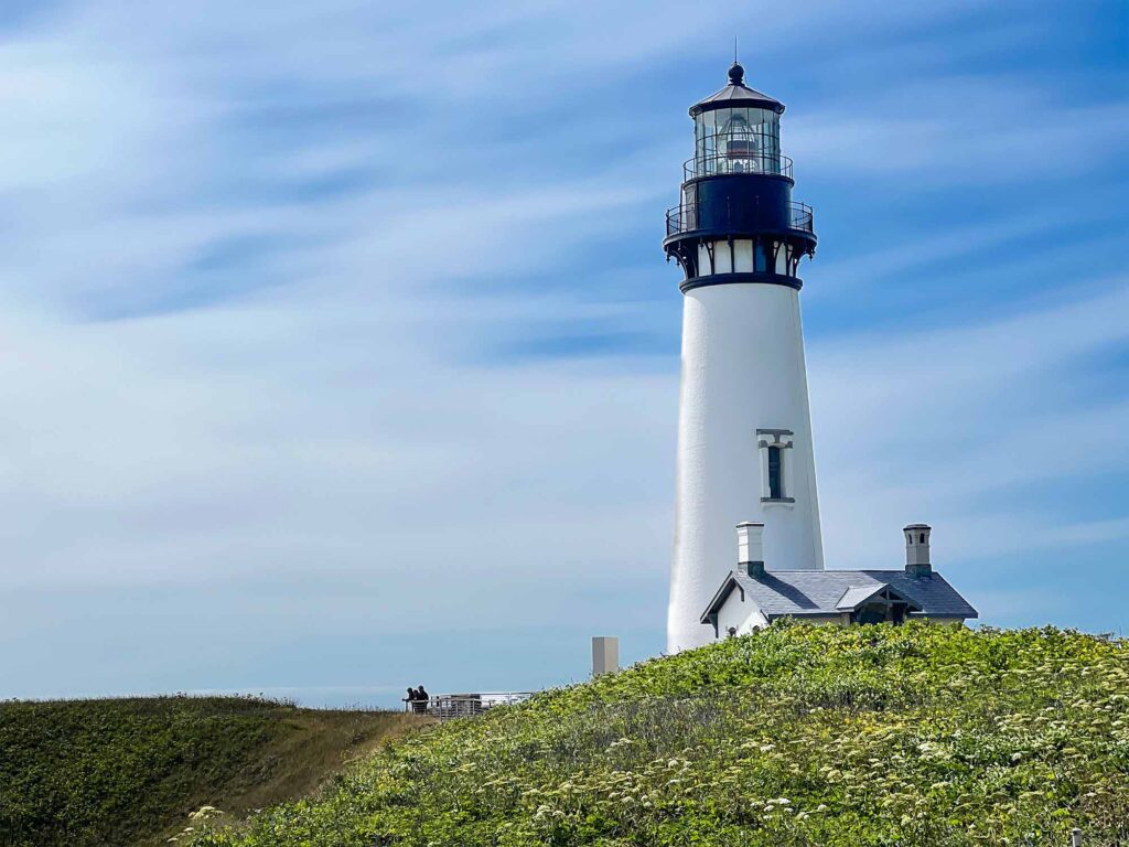 White lighthouse towers over a small white building set against the bright blue sky on the Oregon Coast.