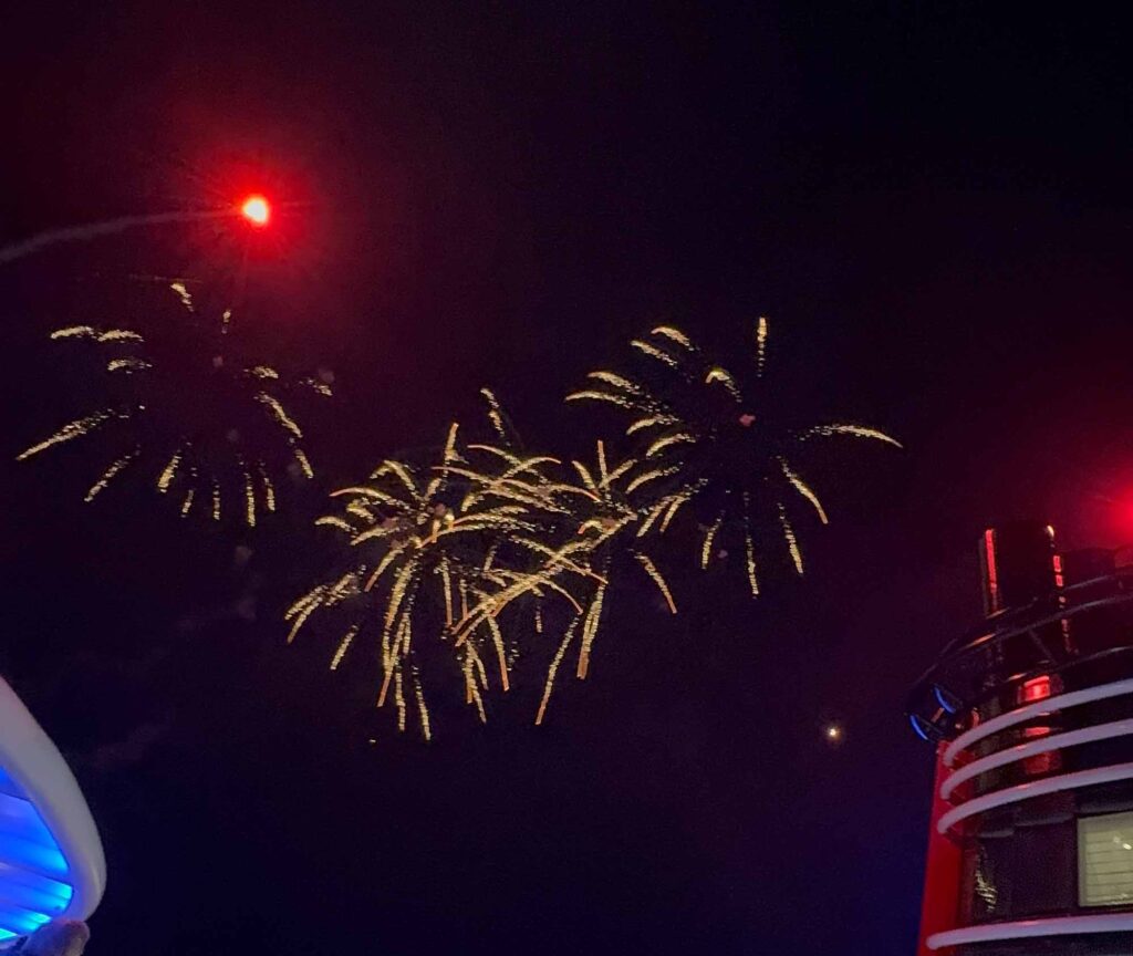 Red and white fireworks erupting over the open ocean as seen from the deck of a disney cruise ship.