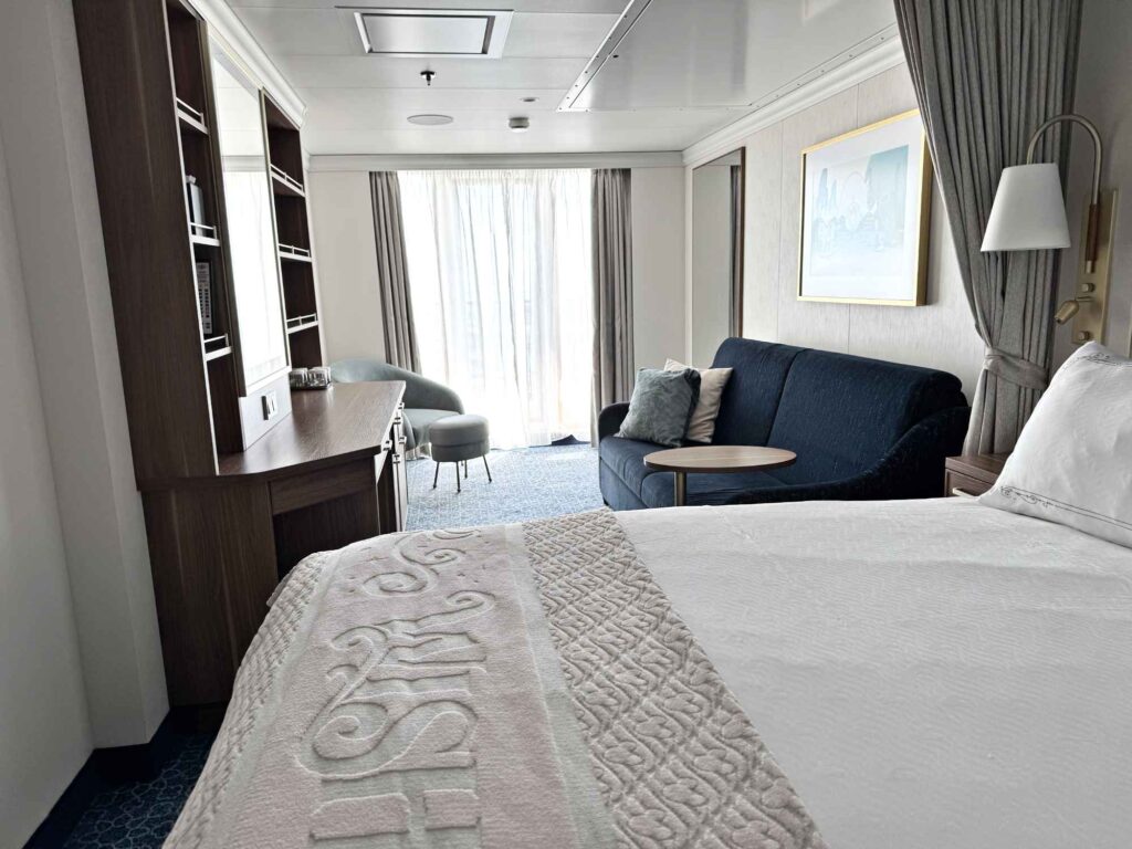 Queen bed with white coverlet before a glass wall with door leading to verandah in a deluxe family stateroom onboard a disney cruise ship.