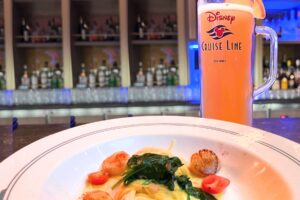 A Disney Cruise beer mug sits behind a plate of scallops and pasta onboard a Disney Cruise ship.